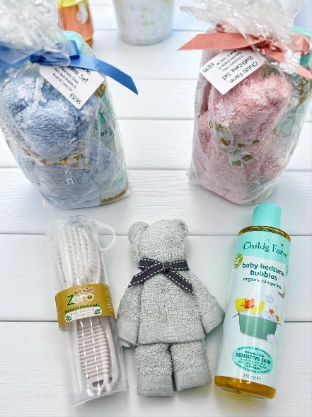 Collection of baby care products gift wrapped and flay lay to show the contents of: a face cloth rolled into the shape of a teddy bear, a eco friendly baby brush and comb set, and a bottle of Childs Farm Baby Bedtime Bubbles on a white wooden surface.