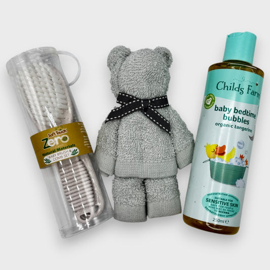 Grey face cloth rolled into the shape of a teddy bear, a baby brush & comb set, and a bottle of 'Childs Farm' baby bedtime bubbles on an off  white background