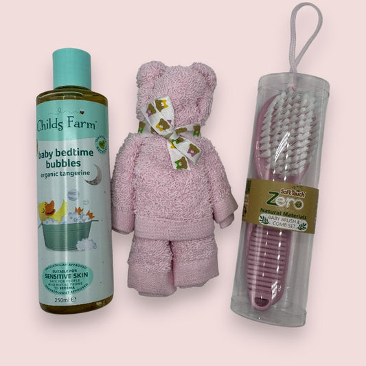 a baby bath gift set from Love & Tiers, which contains: a full sized bottle of Childs Farm Baby Bedtime Bubbles, a Eco friendly Brush & comb set in pink, and a pink face wash cloth rolled into the shape of a teddy bear, on a pale pink background