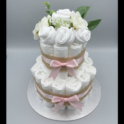 A decorative two-tier nappy cake styled with a baby clothing flower arrangement, with white rolled pampers nappies forming the tiers and the baby clothing forming the roses and carnations, added green leaves, and a hessian and pink ribbon bows on a white cake board against a pale grey background.