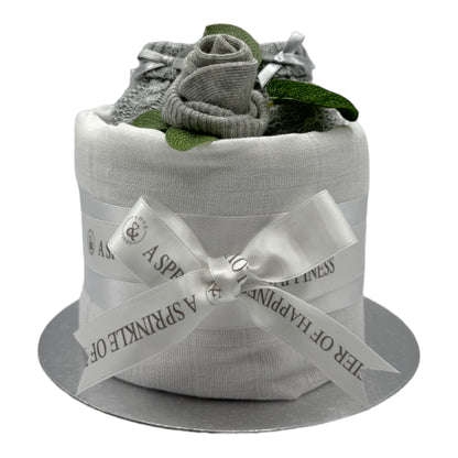 A baby shower nappy cake, wrapped in a white muslin and tied with a white satin ribbon printed with 'A SPINKLE OF HAPPINESS'. The top is decorated with  grey socks rolled into a rosebud, a pair of grey booties, and green leaves, all presented on a silver circular base against a white background.